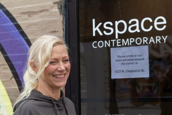 Una mujer rubia de tez clara y sonrisa amplia posa frente a una puerta de cristal sobre la que se lee "kspace contemporary" e indicaciones en inglés sobre la entrada al espacio.
