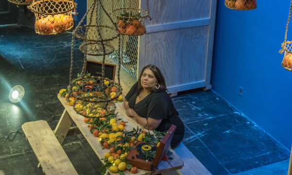 A photograph of playwright and interdisciplinary artist Virginia Grise sitting at a wooden picnic table with fruit on it and in baskets hanging from the ceiling.