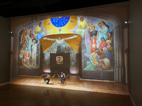 A projection of two musicians playing on a stage in front of Diego Rivera's mural "Creation."