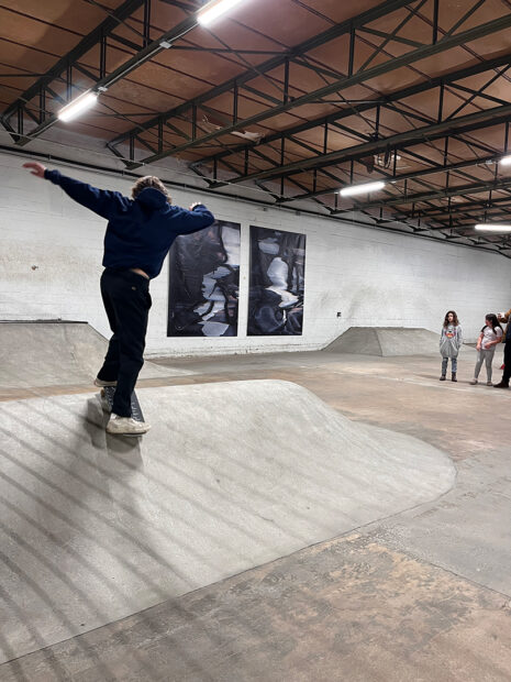 A photograph of a teenager skating in an indoor skatepark with large artworks hanging on the wall.