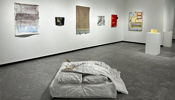 Installation view of works on a wall and a mattress on the floor