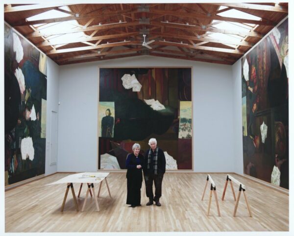 A photograph of Ilya and Emilia Kabakov standing in a gallery with large paintings on each wall.