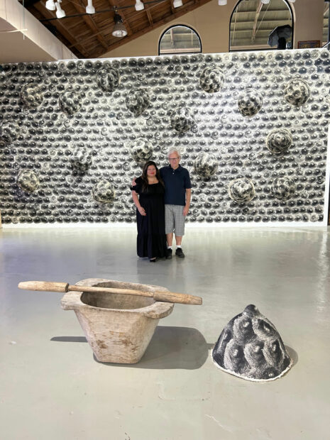 Un hombre y una mujer posan en una galería de paredes blancas frente a un muro cubierto por semicírculos en escala de grises. En primer plano y a la izquierda están una tinaja y una pala de madera y a la derecha una cúpula con un montículo en la cima en escala de grises y pezones impresos en su superficie.