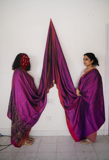 An image of two people standing and facing each other as they share a long traditional Indian cloth. 