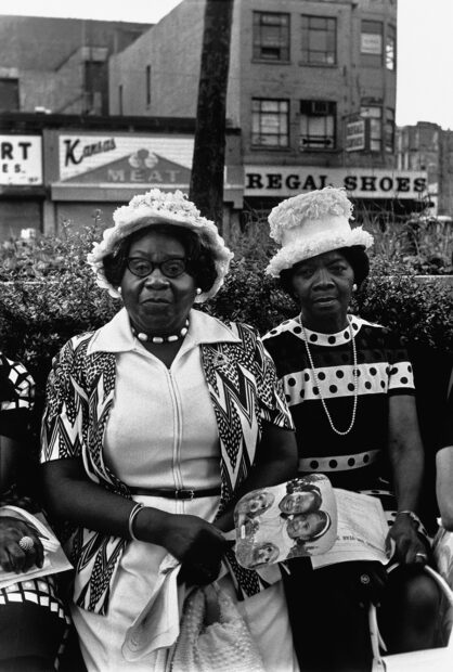 Fotografía en blanco y negro de dos mujeres negras de mediana edad con vestidos y sombreros apropiados para ir a la iglesia. La mujer de la izquierda tiene lentes y sostiene el retrato de dos niñas. Al fondo están un par de negocios con tipografías de la década de los 70 o previas.