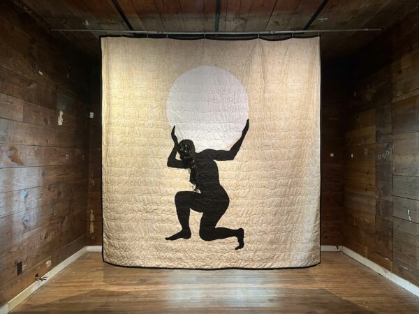 Hanging fabric piece with a woman kneeling and holding white circle on her shoulders