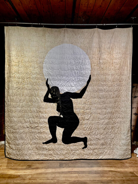 A large quilt by artist Felicia Jordan of a black silhouetted female figure holding the earth on her shoulders.