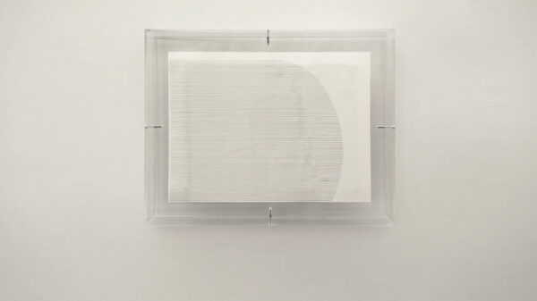Sobre una pared blanca se encuentra una caja de acrílico transparente, dentro de ella delgadas líneas horizontales forman una figura casi rectangular sobre una pieza de papel.