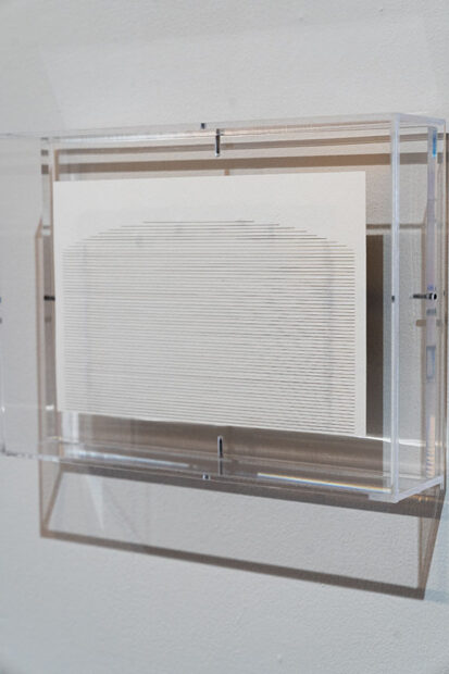 Sobre una pared blanca se encuentra una caja de acrílico transparente, dentro de ella delgadas líneas horizontales forman una figura casi rectangular sobre una pieza de papel.