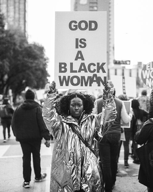 A black and white photograph by Julia Cartwright of a woman holding a protest sign that reads, "God is a Black Woman."