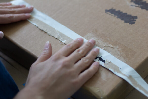Two hands hold a strip of blackout curtain flat against a cardboard box.