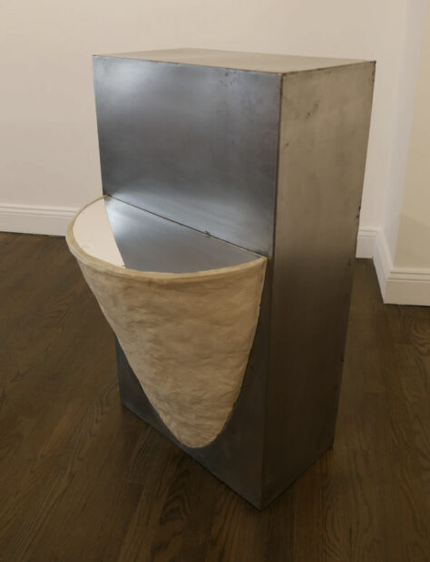 Del costado de un pedestal metálico sobresale una forma parecida a un panal con un corte en la parte superior, donde un espejo refleja la mitad del pedestal.