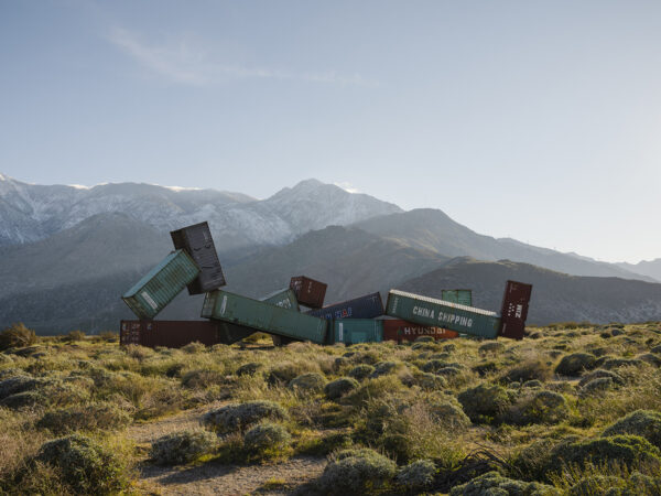 A photograph of a large scale sculpture of a figure constructed from shipping containers with a mountain in the background.
