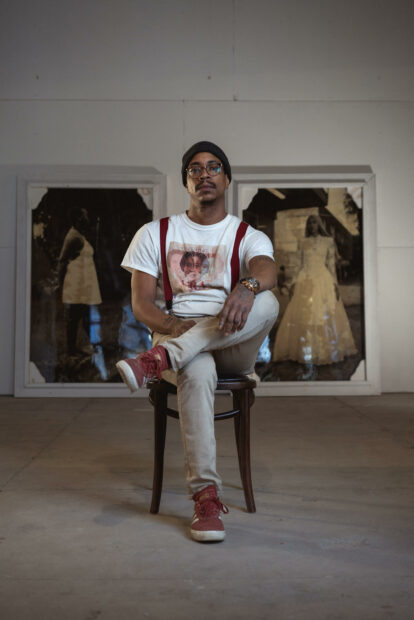 A photograph of artist Colby Deal sitting on a chair with two large sepia photographs behind him.