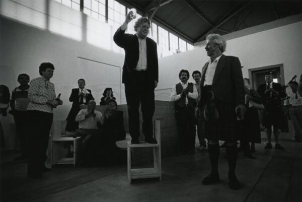 A photograph of Rudi Fuchs standing on a chair and lifting a glass to toast artist Donald Judd.