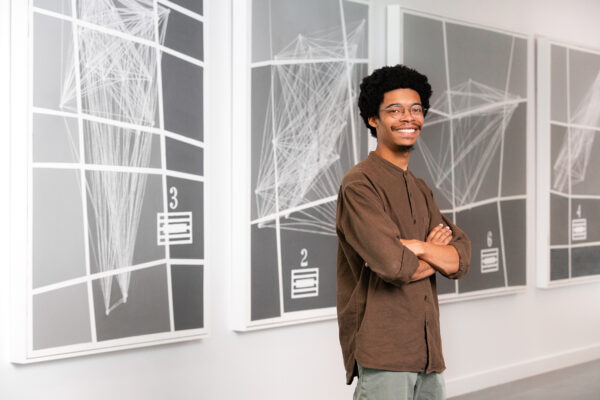 A photograph of artist Corey De'Juan Sherrard Jr. standing in front of his exhibition "Songbook for Black Constellations, for Trio."
