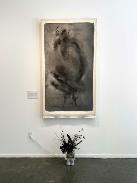 An unstretched drawing on canvas on the wall with a vase of flowers resting on the floor