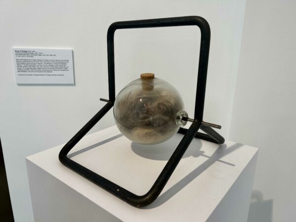 Sculpture of a glass ball with hair inside of it resting on an aluminum armature