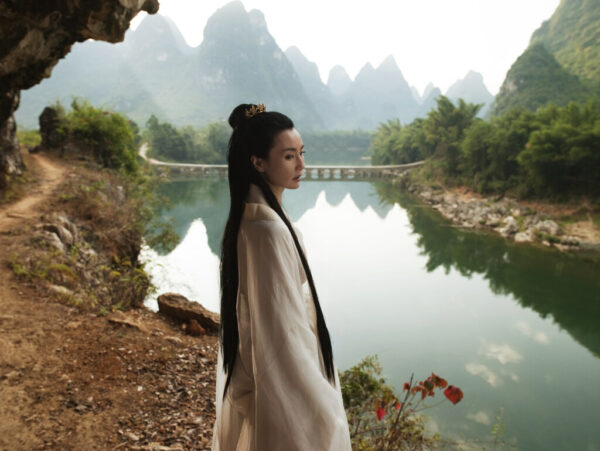 A photograph by Isaac Julien of a woman standing near a river with mountains in the background.