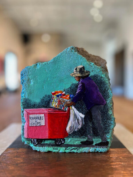 A painting of a person pushing a cart with the a sign that reads "Tamales and Chips." Artwork painted on a stone by Raul Rene Gonzalez.