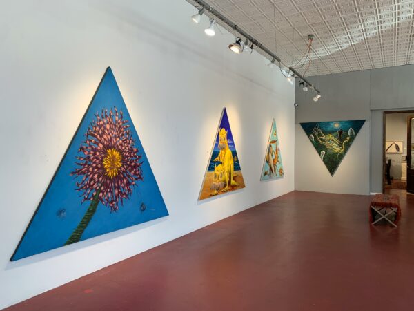 Sobre las paredes blancas de una galería cuelgan varios cuadros triangulares de gran tamaño y distintos colores.