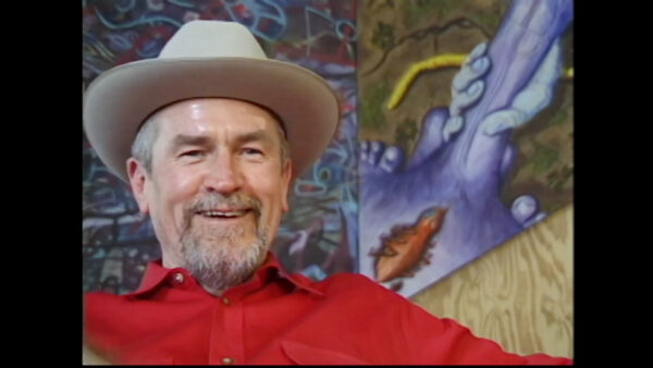A still image from a documentary of a man wearing a red shirt and a cowboy hat.