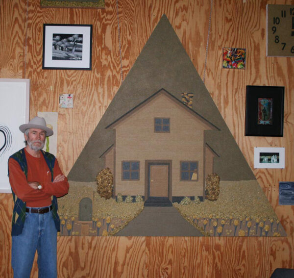 A portrait of a main in his art studio. He stands by a large artwork depicting a house. .