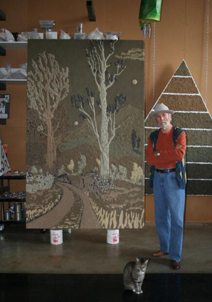 A portrait of a main in his art studio. He stands by a large artwork depicting a road landscape.