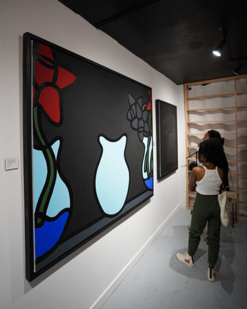 A photograph of gallery visitors looking at two large-scale paintings by Albert Gonzales, featuring simplified flowers in vases.
