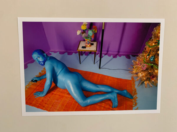 A man, who is painted blue and naked, lies stomach down on an orange rug which is on the ground in a purple-painted room. Behind him is a side table, and to his right is a metallic Christmas tree.