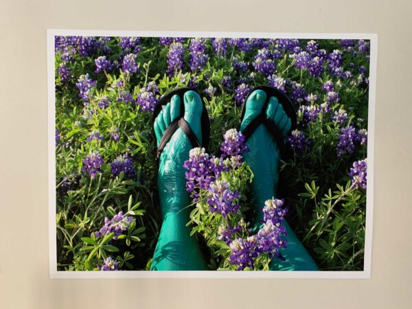 A photograph of green-painted feet wearing black flip flops. The feet are in a field of purple flowers.