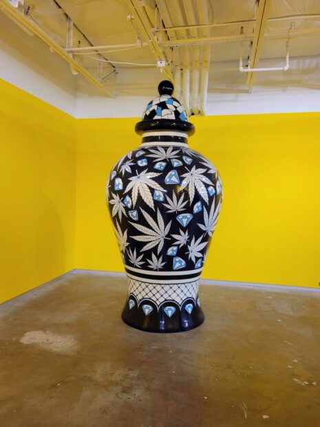 Installation view of a large vase with marijuana leaves against a yellow backdrop