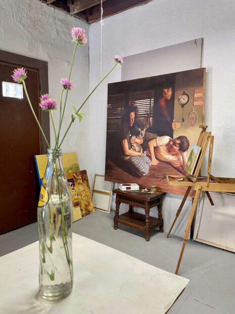 Photo of a studio with flowers in a glass bottle and a figurative painting in the background