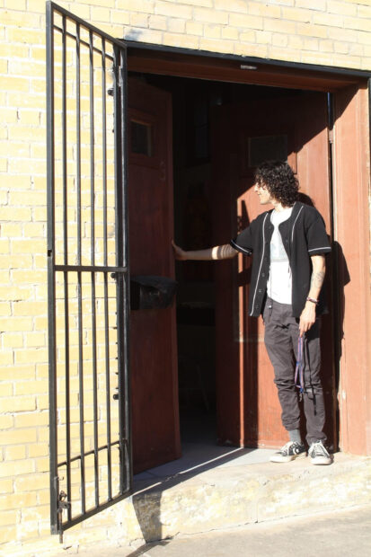 Un hombre de tez apiñonada, cabello rizado y vestimenta casual mira hacia el interior de una construcción de ladrillos mientras sostiene con una mano una puerta de madera.