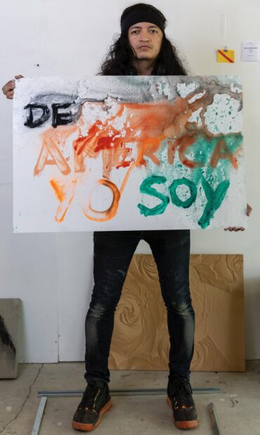A photograph of artist Jesus Benavente with a text based artwork that reads, "De America Yo Soy."
