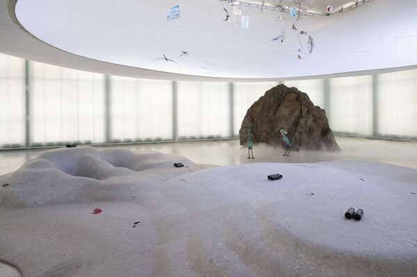 An installation image of a work by Laure Prouvost featuring binoculars scattered on a shore-like creation with sculpted birds standing in the distance.