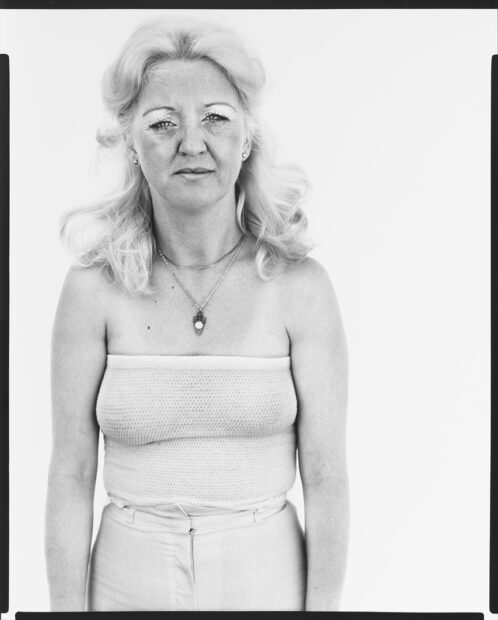 A black and white photograph of a female bartender wearing a partially sheer tube top shirt and white pants..