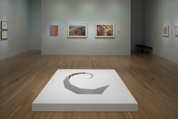 Sobre las paredes grises de una galería cuelgan cuadros de distintos tamaños. En el centro de la sala, una plataforma blanca tiene un diseño de espejos en espiral.