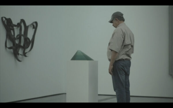 En una galería de paredes blancas, un hombre de edad adulta intermedia, ropa casual y gorra gris observa una escultura piramidal sobre un pedestal.