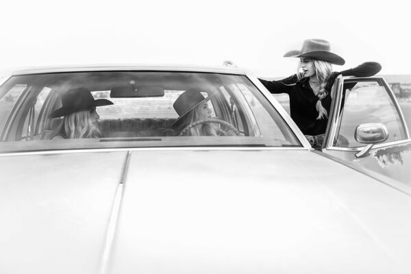 A black and white photograph by Anouk Masson Krantz of three cowgirls, two sitting inside a car and the third standing outside of it.