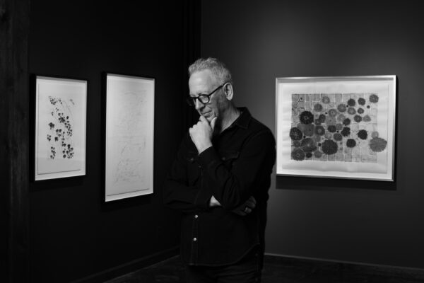 A black and white photograph of a man standing in a gallery in front of artwork.