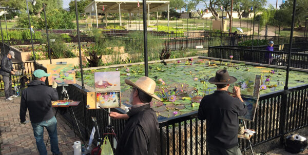 A photograph of a handful of painters with easels painting en plein air near a pond with waterlilies.