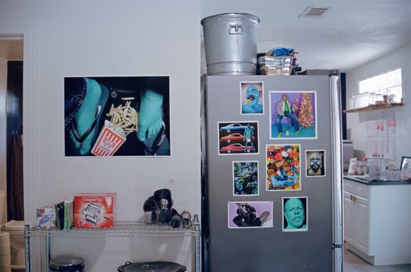Artworks are hung on the side of a refrigerator, and also on a wall, above a shelf.
