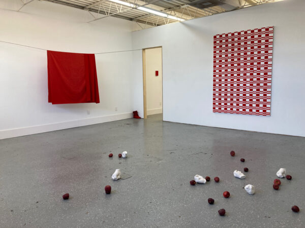 A white-wall gallery contains two large red paintings, with apples and plaster casts of hands strewn on the floor.