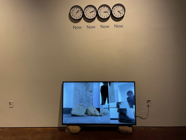 Installation with a flatscreen monitor on two rocks and four clocks with the word "Now" under each