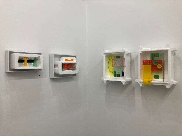 Four artworks consisting of styrofoam and colored plastic in frames are hung in a row at eye level in a gallery
