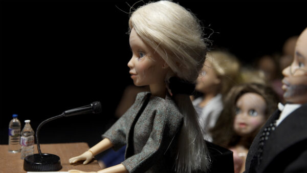 Video still of a doll at a podium in a room full of dolls as an audience