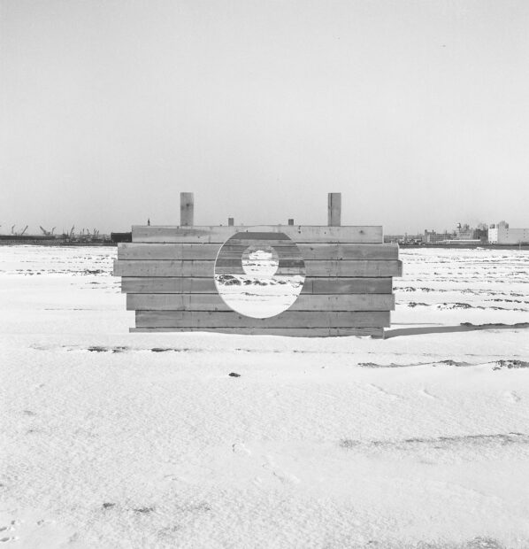Una estructura rectangular de madera tiene un agujero circular en medio. A través de ese agujero, se pueden ver más estructuras parecidas que se extienden sobre el suelo que en esta fotografía en blanco y negro parece nieve.