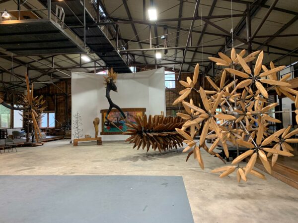 Inside a big warehouse building are multiple wooden sculptures hung from the ceiling.