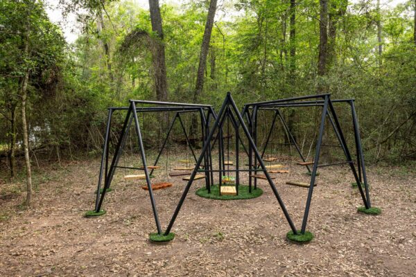 A swingset the looks very spider-like sits in a wooded clearing.